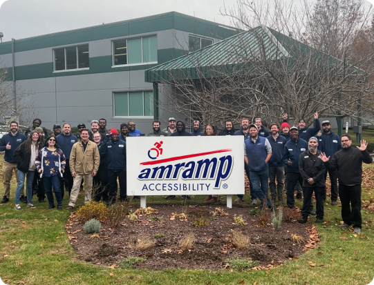 A group photo of the amramp team standing in front of building with a sign with the amramp logo on it